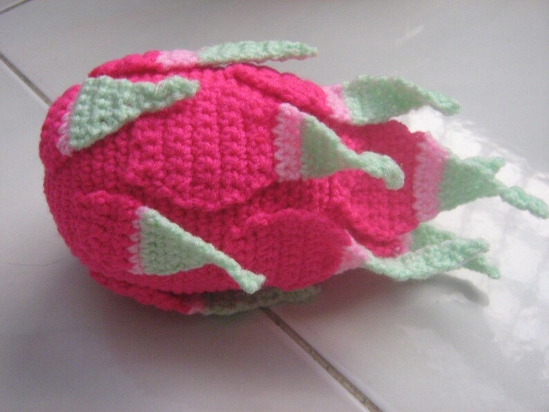 May include: A pink and green crocheted dragon fruit plush toy. The toy is made with a soft yarn and has a realistic shape.