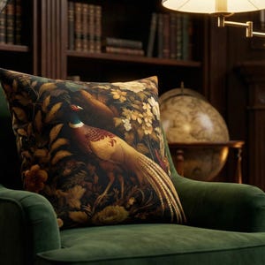 May include: A square throw pillow featuring a detailed print of a pheasant bird surrounded by dark foliage and yellow flowers. The pillow is placed on a dark green velvet armchair. The design includes rich tones of brown, gold, and deep green.