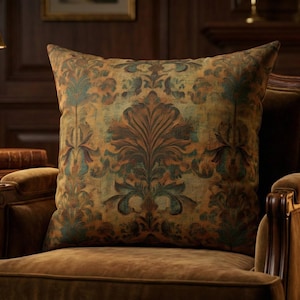 May include: A square throw pillow featuring a dark, muted damask pattern in shades of olive green, burnt orange, and deep teal. The fabric has a textured appearance, and the pillow is positioned upright on a brown upholstered armchair.