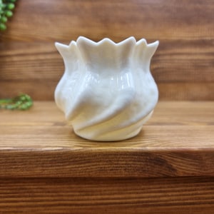 May include: White ceramic planter with a wavy, ruffled design. The planter is sitting on a wooden surface.