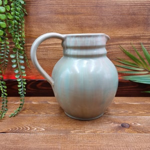 Op de afbeelding: Een lichtgroene keramische kan met een lichtbruine glazuur. De kan heeft een rond lichaam en een handvat.