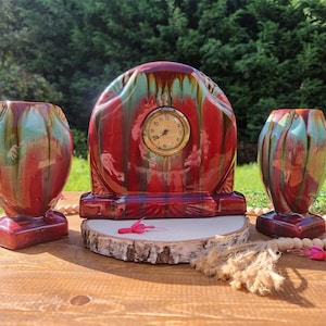 May include: A vintage ceramic clock with a red, green, and brown glaze, flanked by two matching vases. The clock has a round face with gold trim and a cream-colored dial. The vases and clock are on a wooden surface.