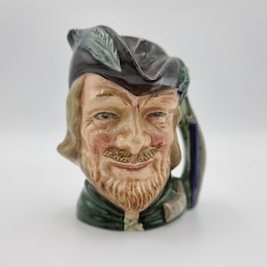 May include: A ceramic mug shaped like a man's head with a green and brown hat and a green and brown jacket. The man has a brown beard and moustache and is smiling.
