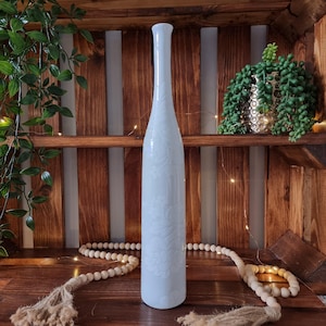 May include: A tall, slender, light blue vase with a floral pattern. The vase is displayed on a wooden shelf with decorative beads and greenery. The vase is approximately 30 cm tall.