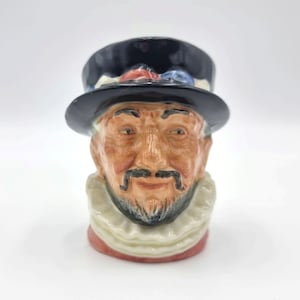 May include: A ceramic figurine of a man's head wearing a black top hat with a red and blue trim. The man has a white ruff collar and a brown beard.