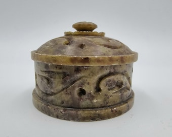 May include: A small, round, stone box with a lid. The box is a mix of brown and tan colors, with carved floral designs around the sides and top. The lid has a small, decorative knob on top.