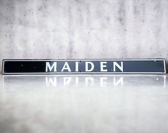 May include: A black and white rectangular sign with the word "MAIDEN" in white letters on a black background. The sign is sitting on a gray surface.