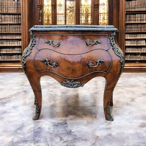 May include: A two-drawer wooden chest with a marble top. The chest has ornate carvings and gold accents. The chest is standing on a marble floor.
