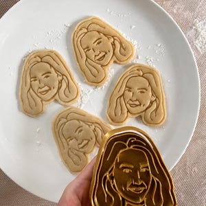 May include: Five light brown cookies, each shaped like a smiling face with long hair, are arranged on a white plate. A gold-coloured cookie cutter, also in the shape of a face, is held in a hand. The cookies are dusted with white powder.