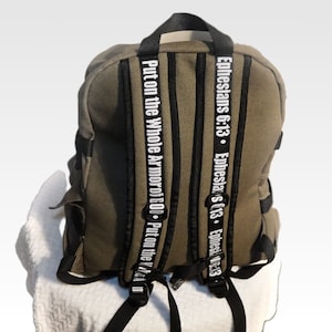 May include: A green canvas backpack with black straps and a white inscription that reads "Ephesians 6:13. Ephesians 6:13. Ephesians 6:13. Put on the Whole Armor of God. Put on the Whole Armor of God."