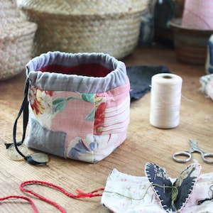 May include: A patchwork fabric drawstring bag with a red interior and gray trim. The bag features floral patterns in pink and red. A spool of white thread, scissors, and a small fabric heart are also visible on a wooden surface.