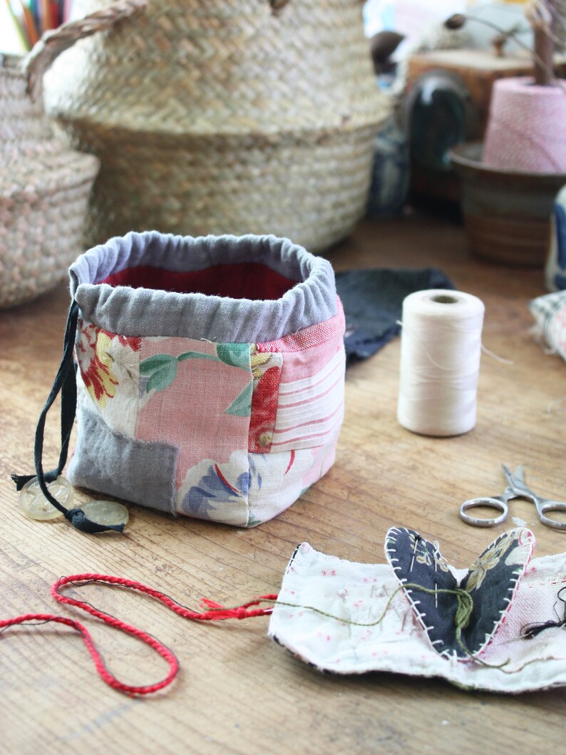May include: A patchwork fabric drawstring bag with a red interior and gray trim. The bag features floral patterns in pink and red. A spool of white thread, scissors, and a small fabric heart are also visible on a wooden surface.