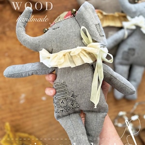 以下が含まれることがあります： 黄色のフリル襟と黄色のリボンが付いたグレーの布製のサーカス象の玩具。象は手作りで、縫製パターンが用意されています。画像には「ANN WOOD handmade」と「circus elephant a sewing pattern」のテキストが表示されています。