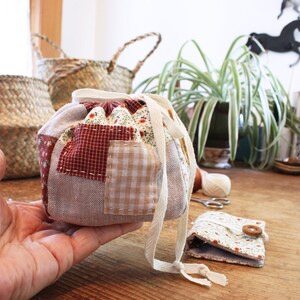 May include: A patchwork fabric drawstring bag with a cream-colored drawstring. The bag features a mix of patterned fabrics in red, beige, and floral prints. A small matching pouch with a wooden button is also visible.