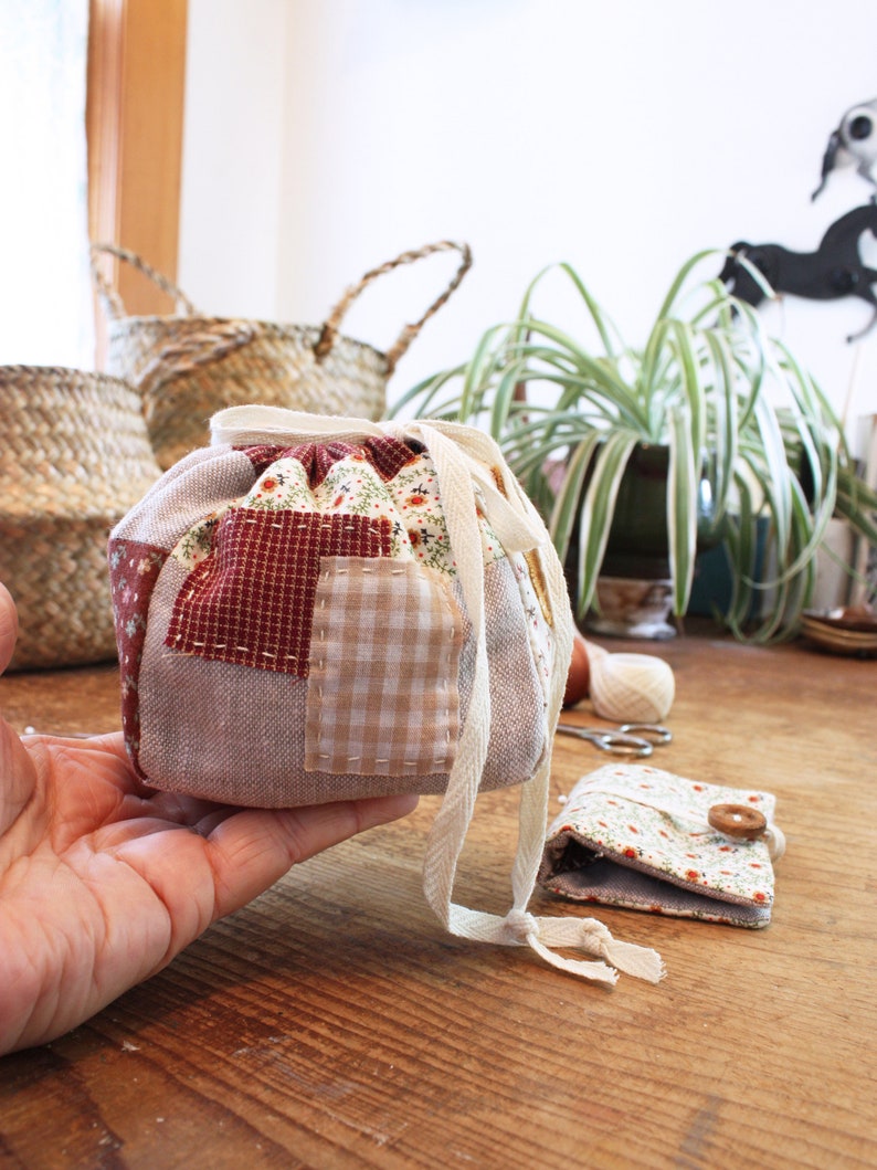 May include: A patchwork fabric drawstring bag with a cream-colored drawstring. The bag features a mix of patterned fabrics in red, beige, and floral prints. A small matching pouch with a wooden button is also visible.
