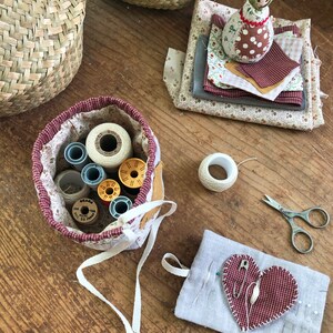 May include: A collection of sewing supplies on a wooden surface. Includes a fabric basket with spools of thread, fabric scraps, a small doll, a heart-shaped pin cushion, scissors, and a roll of twine. The items are in various colors, including red, white, and beige.
