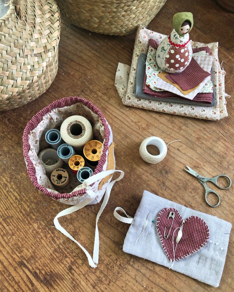 May include: A collection of sewing supplies on a wooden surface. Includes a fabric basket with spools of thread, fabric scraps, a small doll, a heart-shaped pin cushion, scissors, and a roll of twine. The items are in various colors, including red, white, and beige.