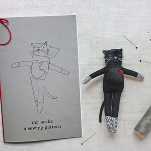 Könnte beinhalten: Eine Schwarz-Weiß-Zeichnung einer Katze mit einem Schal auf einer grauen Karte mit dem Text "mr. socks a sewing pattern".