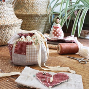 May include: A patchwork drawstring bag with red and floral fabric accents, a heart-shaped pin cushion, and a small doll. The scene includes sewing supplies like thread, scissors, and pins, suggesting a crafting theme. Woven baskets and a houseplant are in the background.