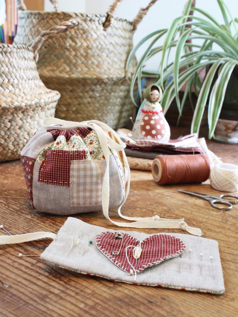 May include: A patchwork drawstring bag with red and floral fabric accents, a heart-shaped pin cushion, and a small doll. The scene includes sewing supplies like thread, scissors, and pins, suggesting a crafting theme. Woven baskets and a houseplant are in the background.