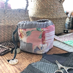 May include: A patchwork fabric drawstring pouch with floral and striped patterns, gray accents, and a gray drawstring. Two woven baskets are in the background. Sewing supplies, including scissors and fabric scraps, are on a wooden surface.