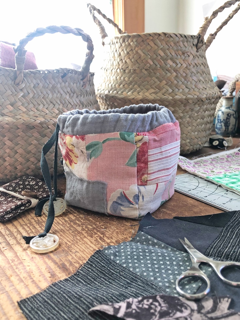 May include: A patchwork fabric drawstring pouch with floral and striped patterns, gray accents, and a gray drawstring. Two woven baskets are in the background. Sewing supplies, including scissors and fabric scraps, are on a wooden surface.