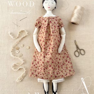 May include: A fabric doll with dark hair in a bun, wearing a tan dress with a small red floral print and white lace-trimmed bloomers. Accessories include a spool of thread, small scissors, sewing needles, buttons, and lace trim. Text reads ANN WOOD handmade and doll clothes a sewing pattern.