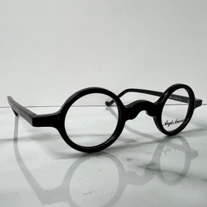 May include: Black round-frame eyeglasses with a keyhole bridge. The glasses have a thick, glossy frame and clear lenses. The text "Anglo American" is printed on the right lens. The glasses are set against a white background.