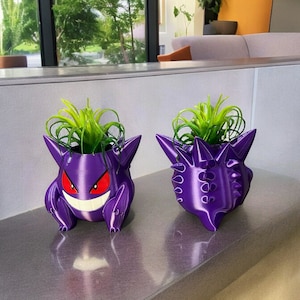 Puede incluir: Dos macetas de Gengar, un Pokémon morado, con plantas verdes en su interior. Las macetas están impresas en 3D y tienen un acabado brillante.