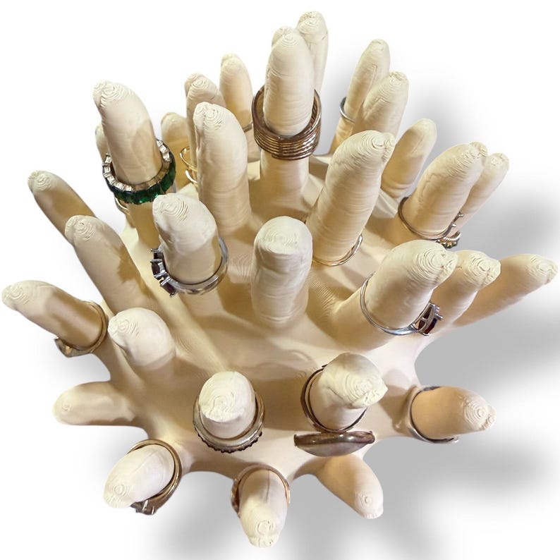 Op de afbeelding: Een cr&egrave;mekleurige ringhouder in de vorm van een hand met meerdere vingers, elk met verschillende ringen. De ringen zijn zilver en goud, sommige met edelstenen. Een decoratief item voor het presenteren van sieraden.