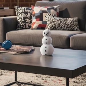 May include: A white snowman figurine with black button eyes and mouth, and an angry expression. The snowman is on a dark gray coffee table in front of a gray sofa with patterned pillows. Blue headphones and magazines are also on the table.