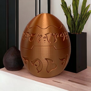 May include: A golden egg-shaped object with intricate carvings and a smooth, metallic finish. The egg is sitting on a wooden surface.