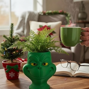 May include: A green Grinch-themed planter with a smiling face, holding a fern plant. A small Christmas tree in a red pot with a bow and snowflake design is to the left. A green mug with steam is held in the background.
