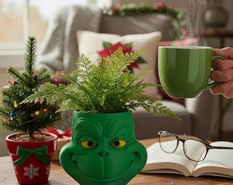 May include: A green Grinch-themed planter with a smiling face, holding a fern plant. A small Christmas tree in a red pot with a bow and snowflake design is to the left. A green mug with steam is held in the background.