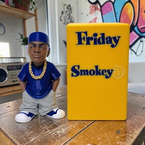 May include: A figurine of a man in a blue shirt, gray pants, and white sneakers with blue accents, wearing a gold chain and a blue cap. Beside it is a yellow block with the words "Friday Smokey" in blue.