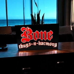 May include: A black sign with red lettering that reads "Bone thugs-n-harmony". The sign is illuminated from behind, creating a glowing effect.