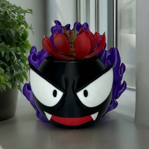 May include: A black and red 3D printed planter shaped like a Pokemon character with purple flames. The planter has a red mouth with white fangs and white eyes. A small red succulent plant is growing in the planter.
