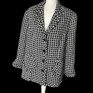 Może przedstawiać: Czarno-biała kraciasta marynarka z frędzlami. Kurtka posiada klasyczny kołnierz, dwie przednie kieszenie i czarne guziki. Tkanina ma teksturowany wygląd, a frędzle dodają dekoracyjny element do projektu.