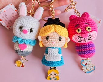 Op de afbeelding: Drie gehaakte sleutelhangers met Alice in Wonderland-personages: een wit konijn met een blauwe en roze jurk, Alice met een blauwe en witte jurk en de Cheshire Cat met een roze en paarse body. Elke sleutelhanger heeft een gouden hanger.