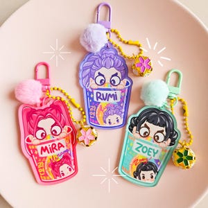 May include: Three keyrings with cartoon characters in ramen cup designs. Each keyring features a different character name: Mira, Rumi, and Zoey. The keyrings have a pom-pom and a decorative charm attached to a chain.