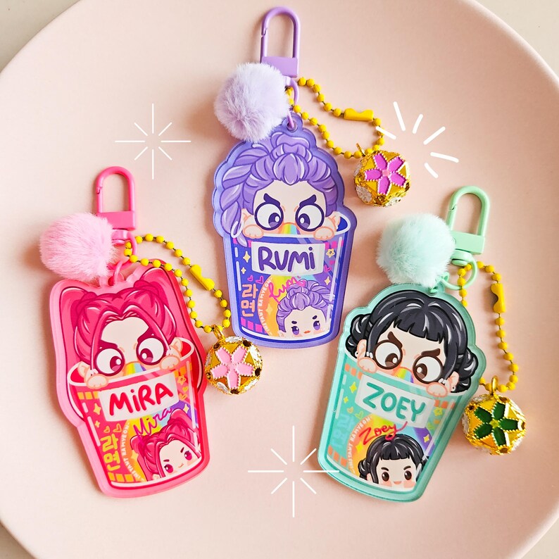 May include: Three keyrings with cartoon characters in ramen cup designs. Each keyring features a different character name: Mira, Rumi, and Zoey. The keyrings have a pom-pom and a decorative charm attached to a chain.