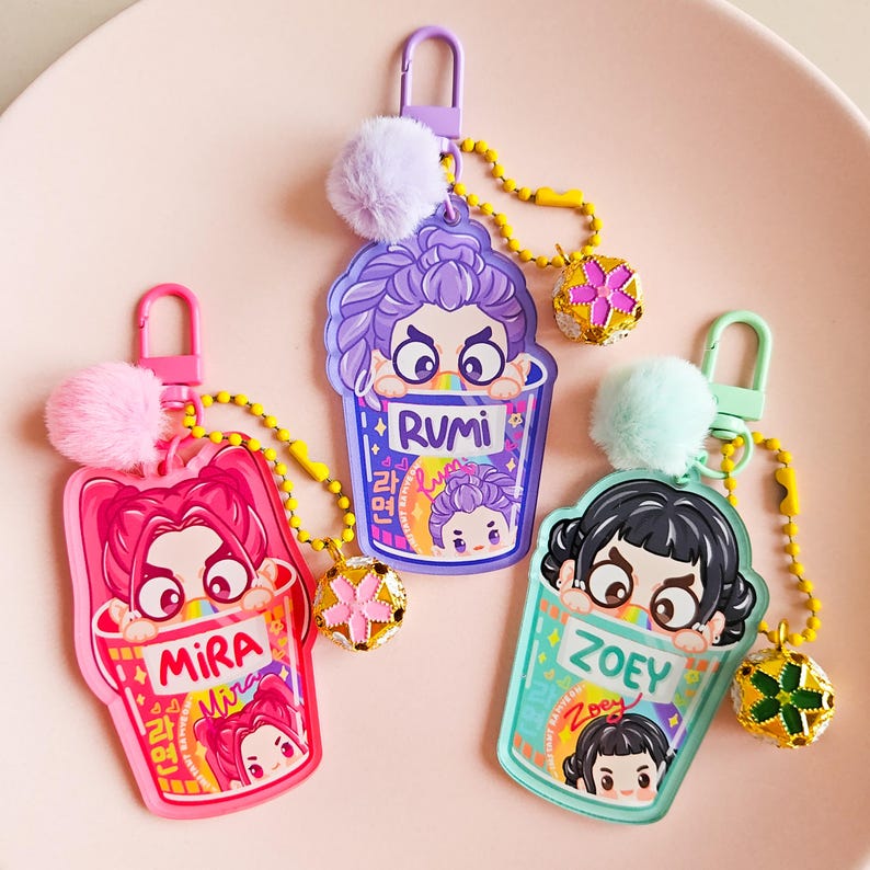 May include: Three keyrings with cartoon illustrations. Each keyring features a character's face peeking out of a cup, with the names "Mira", "Rumi", and "Zoey" displayed. They have a fluffy pom-pom and a decorative charm.