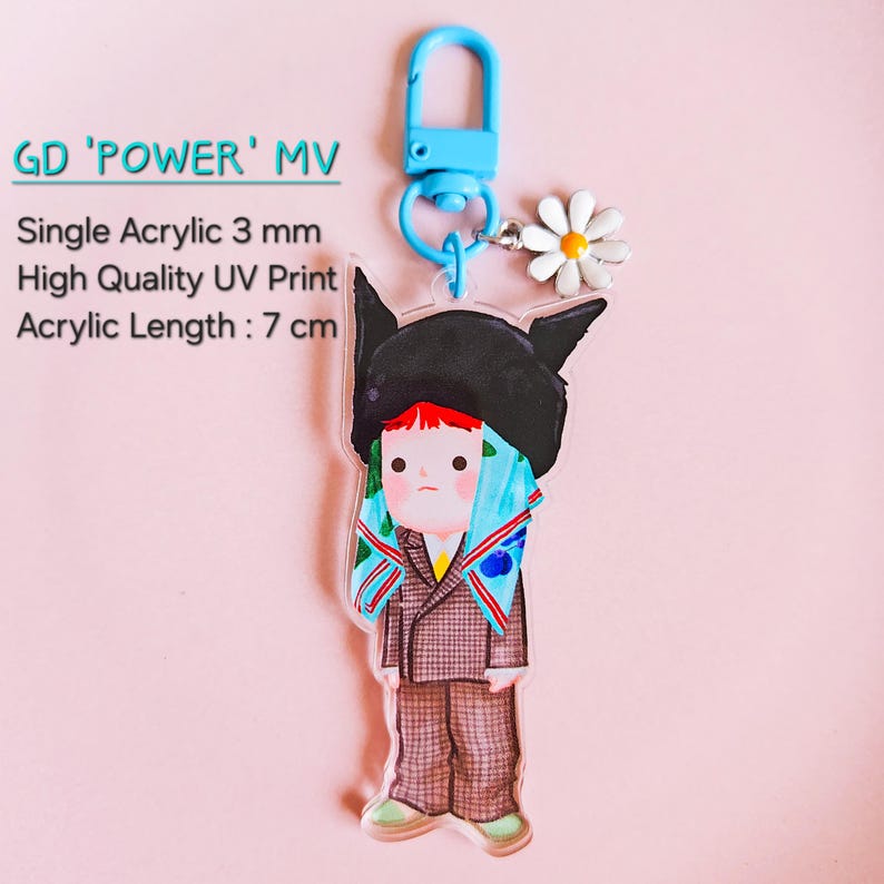 May include: A clear acrylic keychain featuring a cartoon character in a black hat and patterned suit. The keychain has a blue clasp and a small daisy charm. Text on the keychain reads "GD 'POWER' MV" and "Acrylic Length: 7 cm".