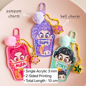 May include: Three acrylic keychains with cartoon character designs, each featuring a pompom and bell charm. The keychains are pink, purple, and teal, with the text "MIRA", "RUMI", and "ZOEY". Total length is 10 cm.