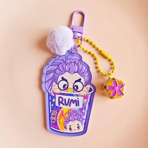 May include: A purple keychain with a cartoon character design emerging from a ramen cup. The cup is labelled "RUMI" with Korean text. A fluffy lavender pom-pom and a gold charm with a pink flower are attached to the keychain.