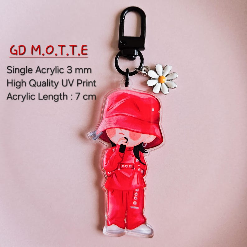 May include: A clear acrylic keychain featuring a cartoon figure in a red outfit and hat. The keychain has a black clasp and a daisy charm. The text "GD M.O.T.T.E" is printed at the top. The acrylic length is 7 cm.