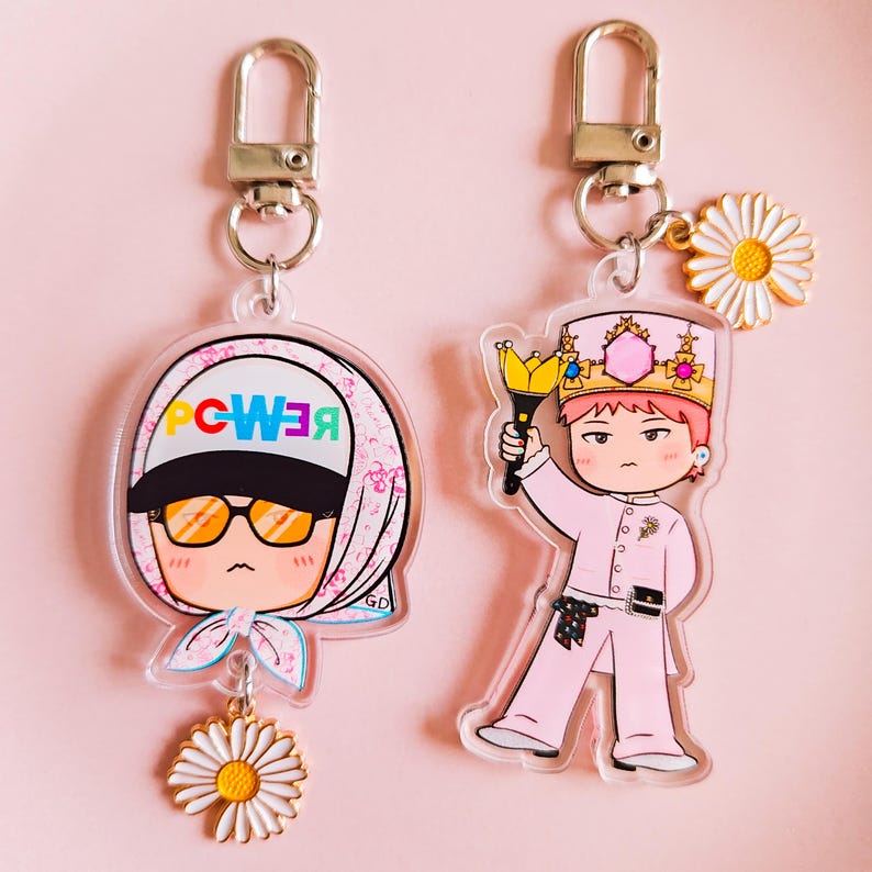 May include: Two acrylic keychains with cartoon illustrations. One features a character wearing sunglasses and a headscarf with the word "POWER" in a circle. The other keychain depicts a character in a pink outfit and crown holding a scepter. Both have daisy charms.