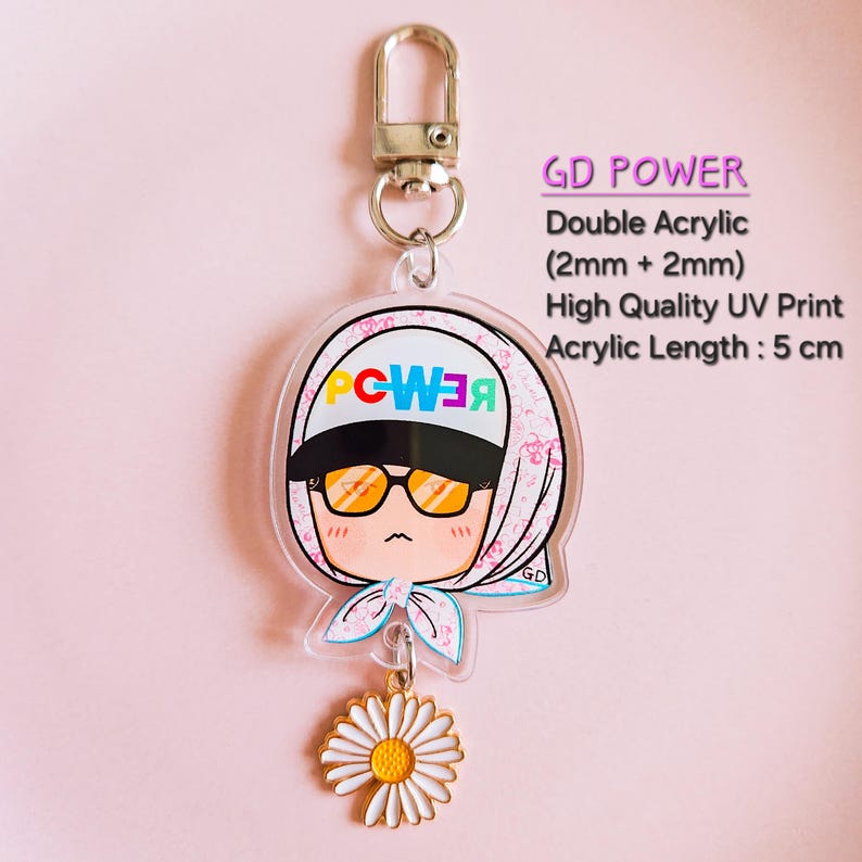 May include: A clear acrylic keychain featuring a cartoon character wearing sunglasses and a headscarf, with the word "POWER" in colorful letters. The keychain has a gold-colored daisy charm and a metal clasp. The acrylic length is 5 cm.