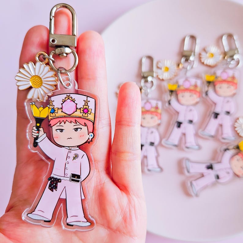 May include: A clear acrylic keychain featuring a cartoon character in a pink outfit and crown, holding a golden scepter. The keychain has a silver clasp and a daisy charm. Additional keychains are visible in the background.