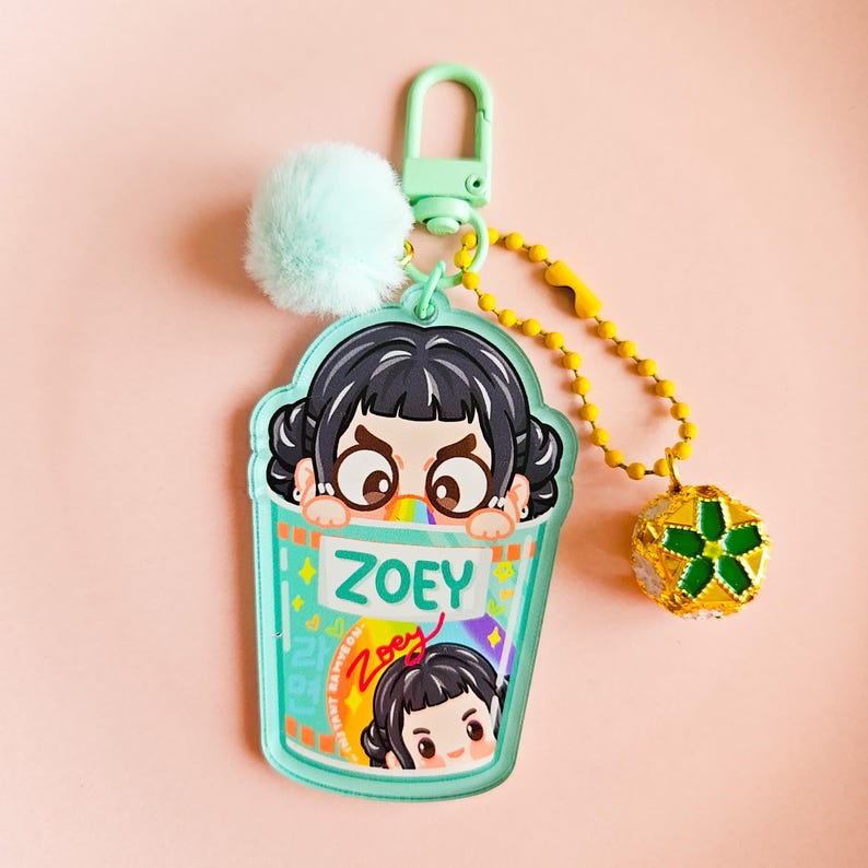 May include: A teal keychain with a cartoon girl design, the name "ZOEY" is printed on the cup. The keychain has a mint green pom-pom, a yellow beaded chain, and a gold and green charm. The background is a light pink.